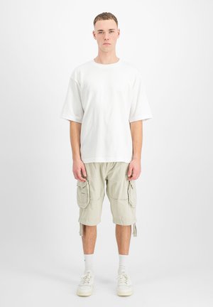 Giovane uomo in piedi rivolto in avanti che indossa una t-shirt bianca oversize, pantaloncini cargo beige, calzini bianchi e scarpe da ginnastica bianche.