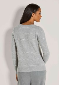 Frau mit langen dunklen Haaren, die einen hellgrauen Strickpullover mit horizontalen Spitzenstreifen und passende graue Hosen trägt, mit dem Rücken zur Kamera.