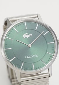 Lacoste käekell rohelise metallist kellapinnaga, hõbedaste aktsentidega, minimalistlike tunni markeritega ja roostevabast terasest rihmaga. Kellal on logo 12. kohal.