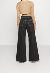 Donna che indossa pantaloni neri a gamba larga con vita alta e cuciture bianche, insieme a un maglione a maglia corto color sabbia, in piedi su uno sfondo semplice.