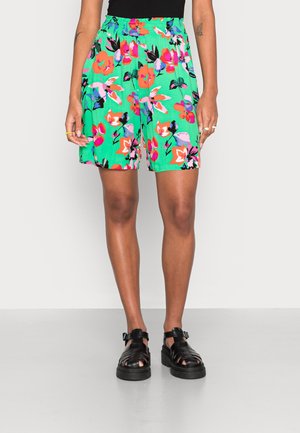 Vrouw draagt groene shorts met elastische taille en een felroze en paarse bloemenprint, gecombineerd met zwarte sandalen met dikke bandjes.
