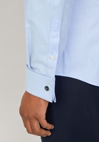 Camicia a quadri blu chiaro con colletto a punta, polsino con bottone singolo e bottone di accento tondo navy. Tessuto in cotone liscio.