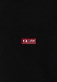 Camiseta de algodón negra con un parche rectangular bordado en rojo que lleva la palabra "GUESS" en letras blancas cerca del centro.