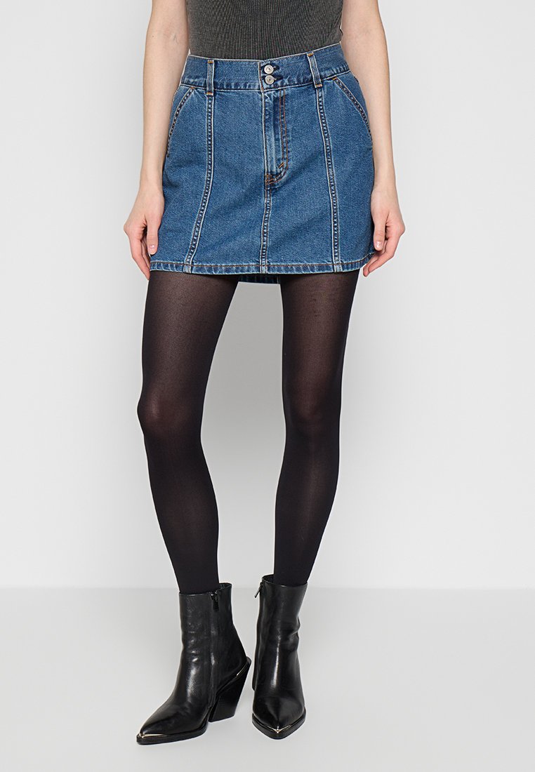 Denim mini skirt in blue with five pockets, button details, and a structured waist. Worn with black tights and ankle boots.