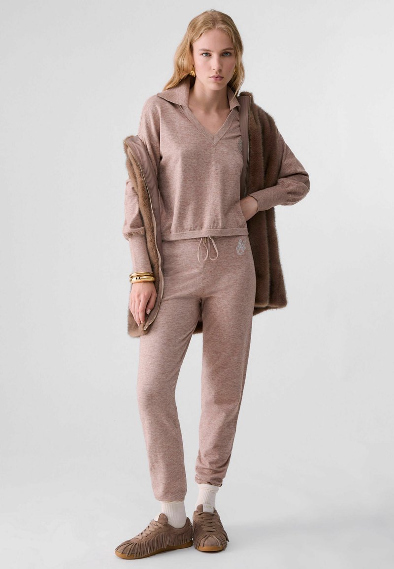Femme portant un survêtement en maille marron, une veste marron drapée sur les épaules, des bracelets dorés et des baskets marron à franges avec des chaussettes blanches.