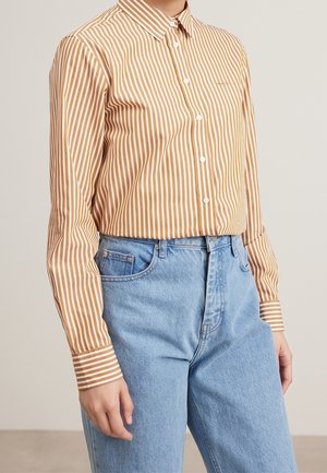 Personne portant une chemise à boutons rayée verticalement marron et blanche, rentrée dans un jean taille haute bleu clair, sur un fond uni.