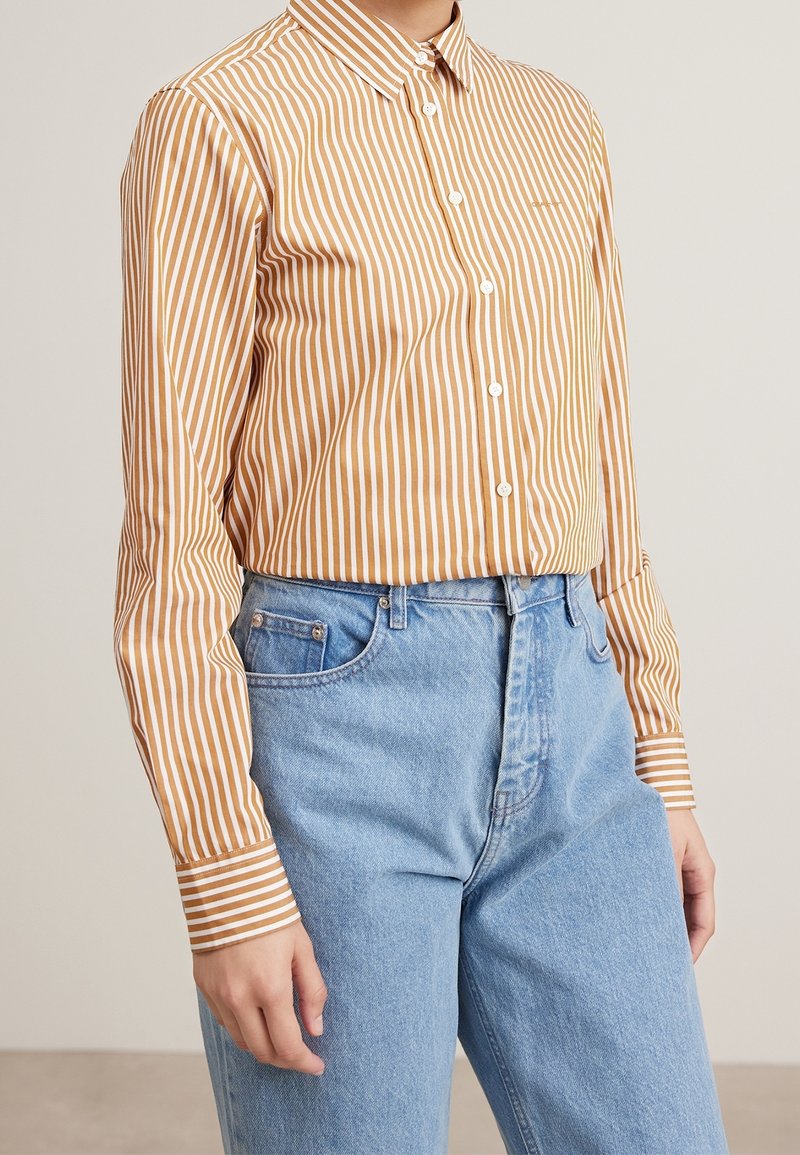 Personne portant une chemise à boutons rayée verticalement marron et blanche, rentrée dans un jean taille haute bleu clair, sur un fond uni.