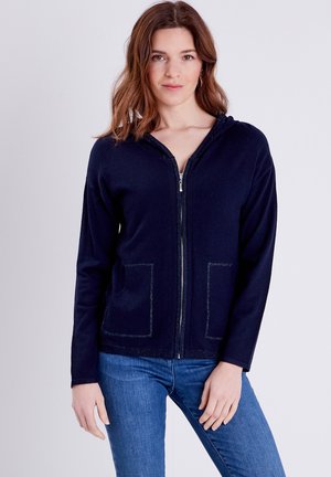 Marine blauwe rits-hoodie van zachte stof met voorzakken, voorzien van contrasterende stiksels en een capuchon. Model heeft het gecombineerd met blauwe jeans.