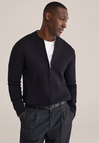 Tamsiai mėlynas megztinis su užtrauktuku, stovinčiu apykakliu ir priekinėmis kišenėmis, derinamas su pilkomis siaurai čiuožiančiomis kelnėmis. Po apačia dėvimas baltas marškinėlis.