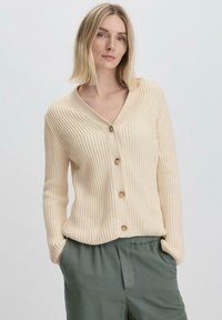 Beige Rippstrick-Cardigan mit tiefem V-Ausschnitt, fünf Holzknöpfen auf der Vorderseite und langen Ärmeln. Getragen über grünen, lässig geschnittenen Hosen.
