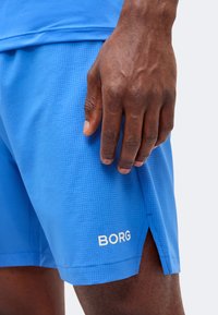 Χέρι με σκουρόχρωμο δέρμα που ακουμπά κοντά στον μηρό, φοράει μπλε σορτς Borg με μικρή πλευρική σχισμή και σχέδιο πλέγματος.