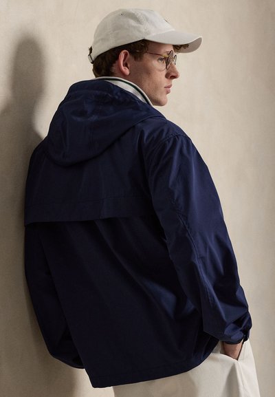 Jeune homme portant des lunettes et une casquette blanche, porte une veste à capuche bleu marine, de profil contre un mur beige uni, une main dans la poche.