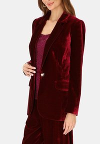 Bordowa aksamitna marynarka z pojedynczym ozdobnym guzikiem, klapami z wycięciem oraz dwoma przednimi kieszeniami, noszona na wzorzystej śliwkowej bluzce.