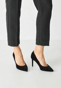 Décolleté neri in suede con design a punta e tacco stiletto di 10 cm, abbinati a pantaloni neri su misura. Texture liscia, accenti minimali.