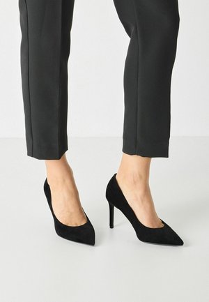 Décolleté neri in suede con design a punta e tacco stiletto di 10 cm, abbinati a pantaloni neri su misura. Texture liscia, accenti minimali.
