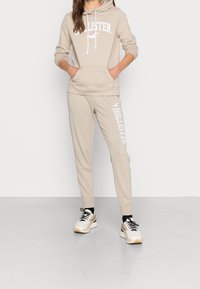 Žena v béžové mikině Hollister a odpovídajících teplákách s bílým logem, černými ponožkami a bílými teniskami, stojí se založenýma rukama v kapsách.