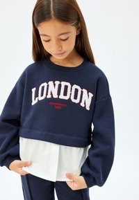 Sudadera recortada azul marino con "LONDRES" bordado en blanco y acentos rojos, superpuesta sobre un dobladillo de camisa blanca texturizada. Mangas largas y abullonadas.