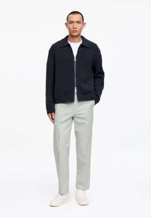 Man wearing a black zip-up cardigan, white t-shirt, light gray trousers, and white sneakers, standing with one hand in pocket.