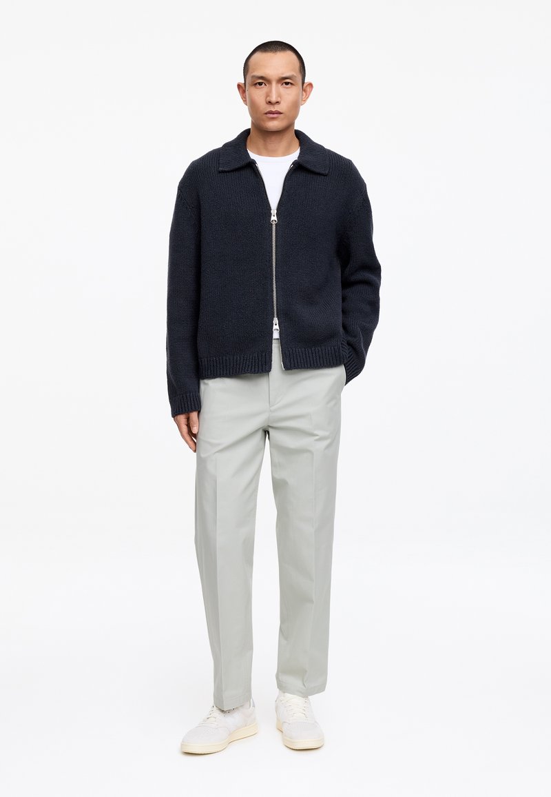 Homme portant un cardigan noir zippé, un t-shirt blanc, un pantalon gris clair et des baskets blanches, debout avec une main dans la poche.
