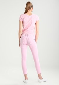 Žena s culíkem, oblečená v světle růžovém tričku s krátkým rukávem a odpovídajících růžových kalhotách, stojící s otočením zády k jednobarevnému bílému pozadí.