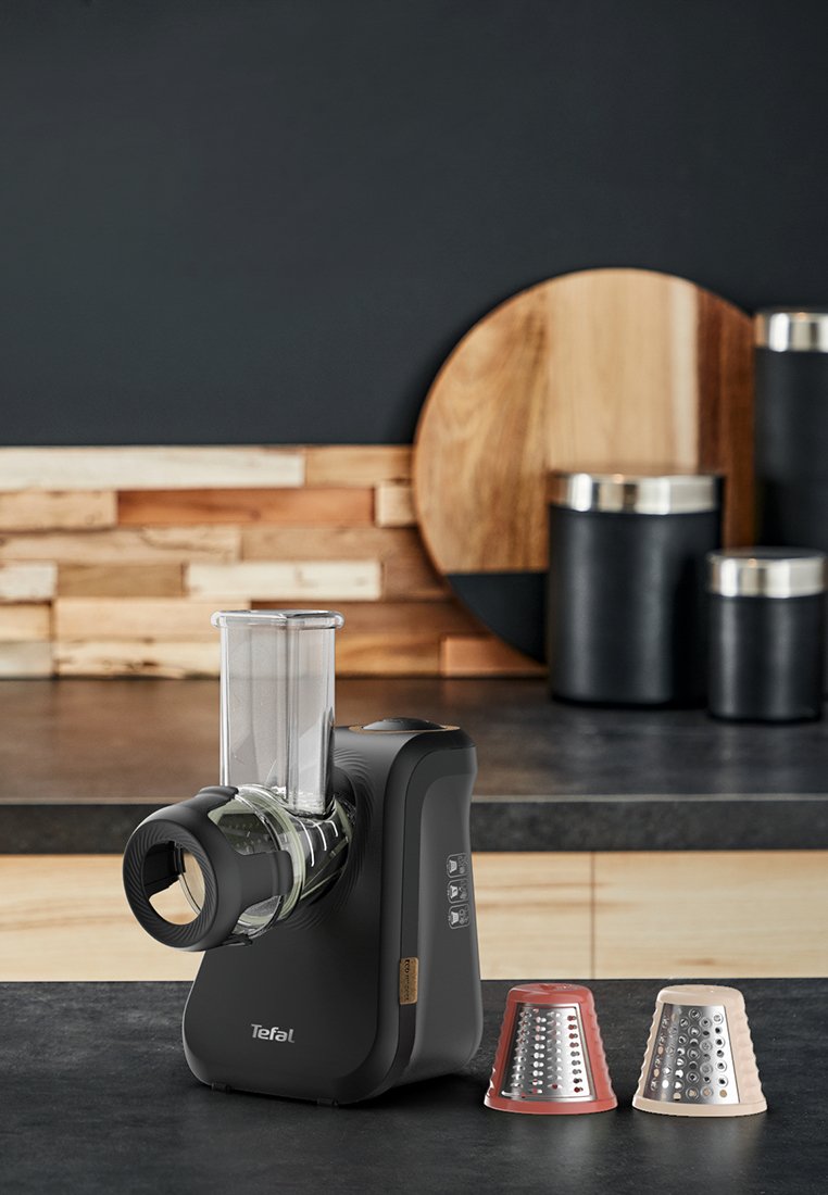 Robot culinaire électrique noir avec un récipient transparent, trois accessoires de râpage interchangeables et un design élégant. Boutons notables sur le côté.