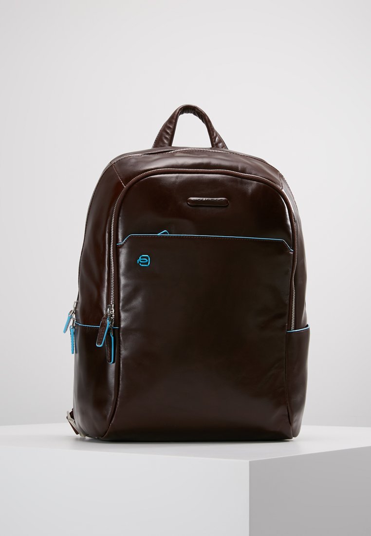 Mochila de cuero marrón con dos bolsillos frontales con cremallera, detalles en azul, asa acolchada y correas de hombro ajustables. Textura suave, diseño minimalista.