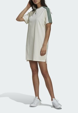 Vrouw draagt een witte Adidas T-shirtjurk met groene strepen op de mouwen en witte Adidas sneakers, staat met één hand bij haar hoofd.