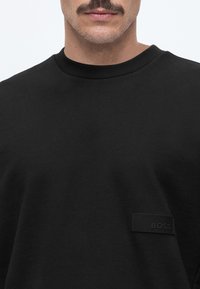 Homme avec moustache portant un t-shirt noir uni à col rond avec un petit patch noir rectangulaire étiqueté "BOSS" sur la poitrine.