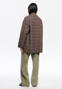Chaqueta de cuadros de gran tamaño en marrón y beige, con un corte relajado. Combinada con pantalones verdes holgados y calzado texturizado.