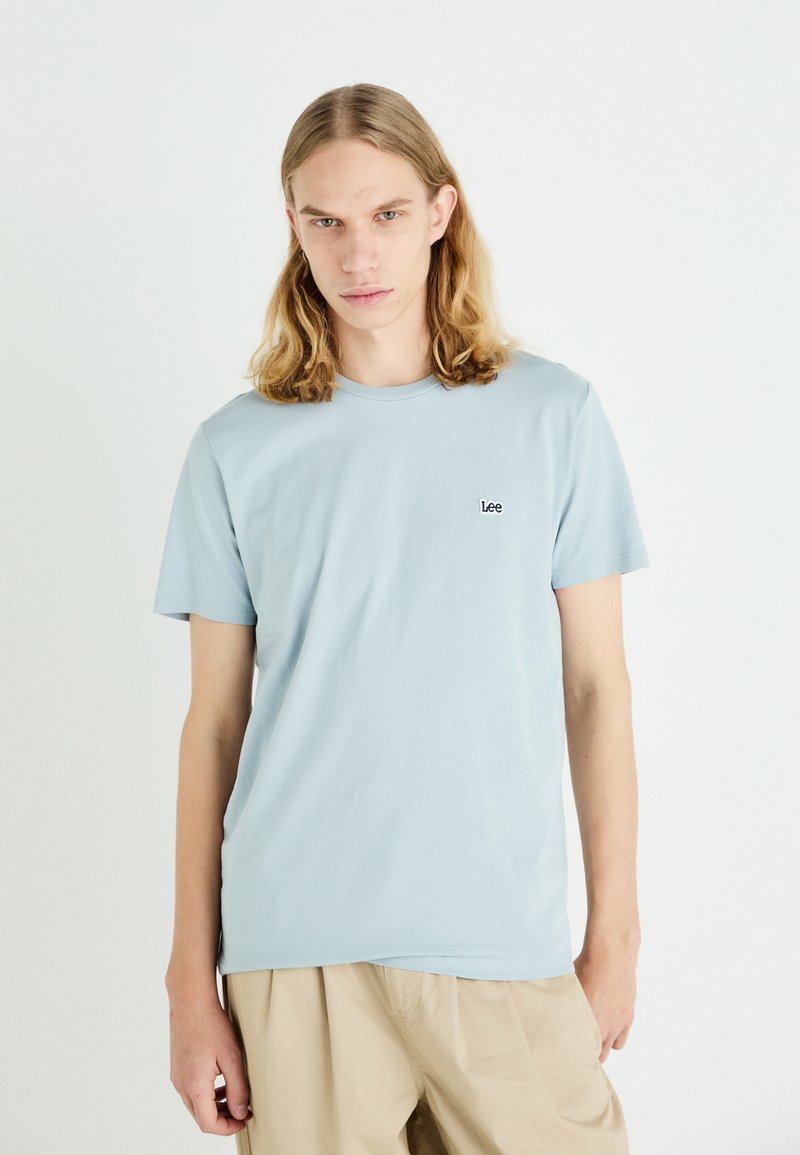 Camiseta de algodón azul claro con cuello redondo y mangas cortas. Presenta un pequeño logotipo negro de "Lee" en el pecho. Textura suave, corte relajado.