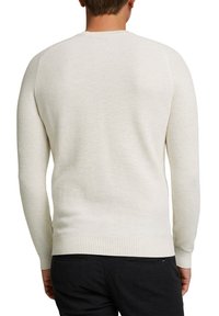 Homme portant un pull beige texturé à manches longues et un pantalon noir, vu de dos sur fond blanc.