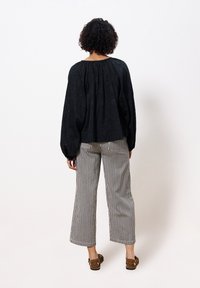 Zwarte blouse met structuur en brede mouwen, gecombineerd met gestreepte, wijde broek in zwart en wit. Model draagt bruine platte sandalen.
