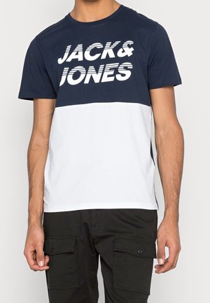 Mężczyzna noszący granatowo-biały t-shirt z napisem "JACK & JONES" drukowanym grubymi białymi literami na piersi.
