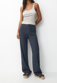 Kobieta w beżowym topie na ramiączkach, granatowych szerokich spodniach i ciemnobrązowych sandałach na platformie, stojąca przy białej ścianie.