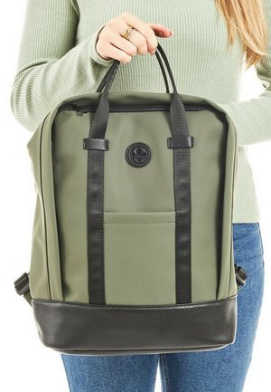 Sac à dos en tissu vert avec des bretelles et un fond noirs. Il comprend une poche frontale et un logo rond. Texture lisse avec des détails en contraste.