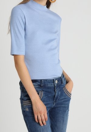 Femme portant un haut bleu clair à manches courtes avec col montant et un jean en denim bleu orné, avec une main dans la poche.