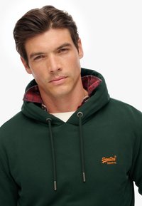 Donkergroene hoodie met een trekkoord, voorzien van een oranje logo op de borst en een geruite rood- en zwartgekleurde flanellen voering in de kraag.