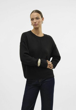 Maglione nero lavorato a maglia con scollo tondo, vestibilità rilassata e maniche lunghe. I dettagli degni di nota includono un tessuto texture e un bracciale decorativo.