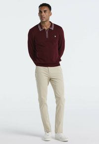 Burgunderfarbener Pullover mit weißem Kragen und Besatz, kombiniert mit beigen Hosen und weißen Sneakers, getragen von einer stehenden männlichen Figur vor einem hellen Hintergrund.