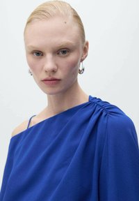 Abito azzurro a spalla unica realizzato in tessuto morbido con dettagli arricciati sulla spalla. La modella indossa grandi orecchini a cerchio e ha i capelli pettinati all'indietro.