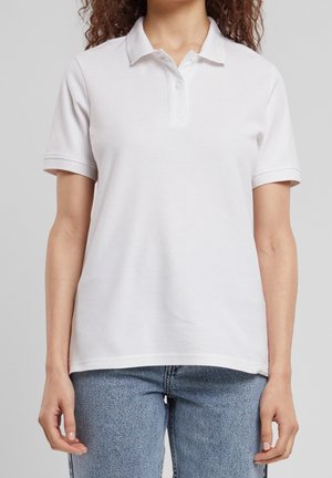 Vrouw draagt een eenvoudige witte polo met korte mouwen, niet in de broek gestopt, gecombineerd met lichtblauwe jeans, tegen een lichtgrijze achtergrond.