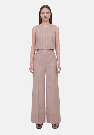 Donna in piedi rivolta verso di noi, indossa un top beige senza maniche a righe sottili e pantaloni larghi abbinati con scarpe marroni a righe.