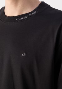 T-shirt nera in cotone con collo giro, caratterizzata da un piccolo logo grigio "ck" sul petto e dal nome del marchio ricamato a contrasto sul colletto.
