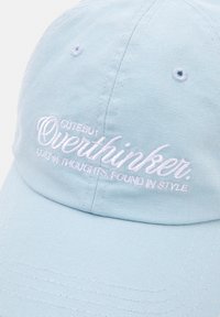 Light blue cap made of textured fabric; features white embroidered text reading "Cute but Overthinker" with additional text below.