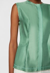 Haut sans manches vert menthe avec ourlets bruts et détails de couture verticale porté par une personne à la peau mate.