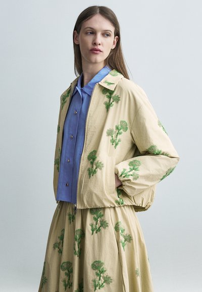 Femme portant une veste beige et une jupe avec une broderie florale verte, chemise à boutons bleue, debout devant un fond gris clair.