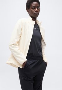 Άτομο που φοράει εκρού μπουφάν πάνω από μαύρο πουκάμισο Salomon και μαύρο παντελόνι, στέκεται με τα χέρια στις τσέπες μπροστά σε απλό φόντο.