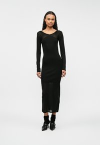 Czarna sukienka z długim rękawem, dopasowaną górą i przezroczystymi detalami przy dekolcie; ma długość do połowy łydki i boczne rozcięcia.