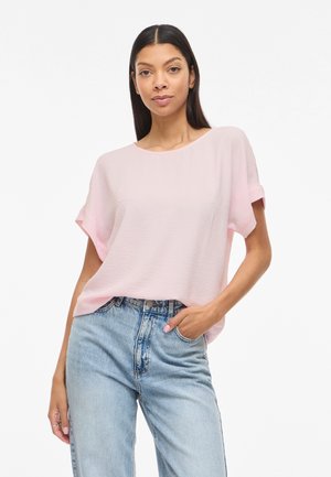 Kvinde stående iført en lyserød kortærmet bluse stoppet ind i højttaljede lyseblå jeans, den ene hånd i lommen, neutral baggrund.