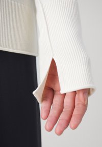 Top a maniche lunghe bianco a coste con polsini aperti, che rivelano una mano. Il tessuto appare morbido con una finitura testurizzata, abbinato a un fondo scuro.